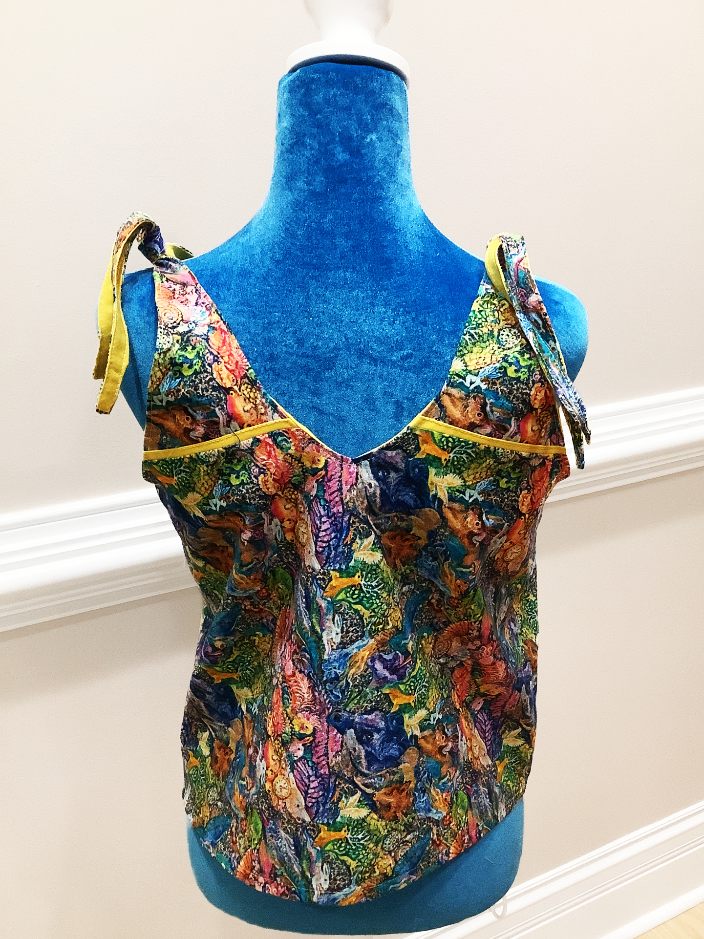 Back view of a colorful tank top on a blue manniquin with a beige background. The tank top has tied straps at the shoulders.