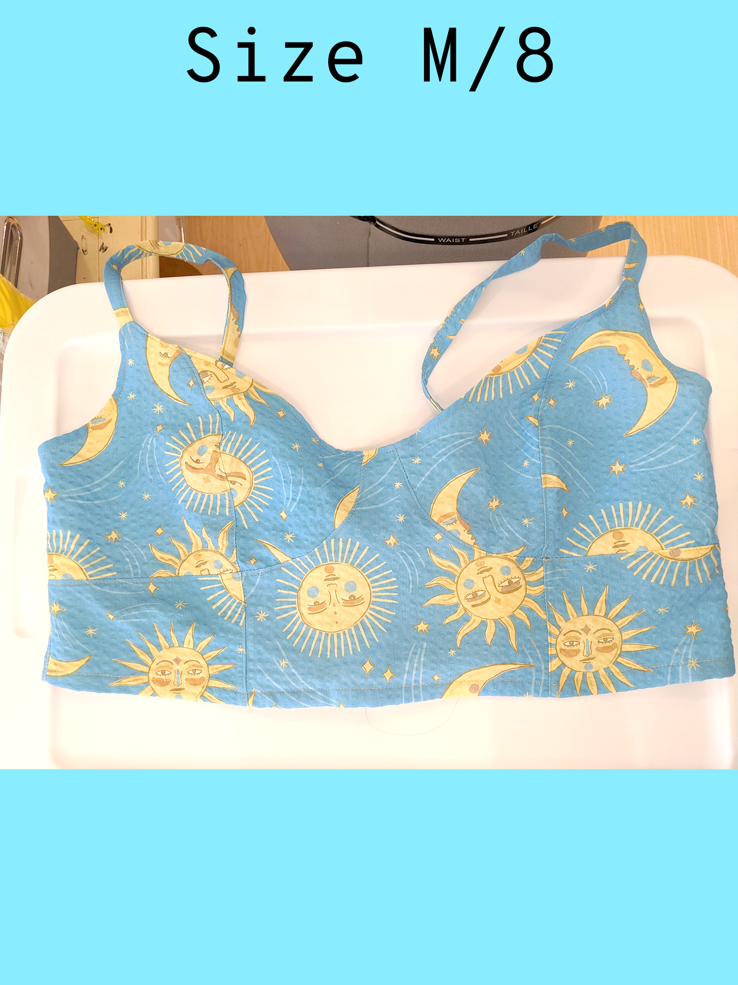 A light blue cropped tank top with a yellow sun and moon print and gold topstitching, displayed flat on a white surface. With a text overlay that reads, "Size M/8".