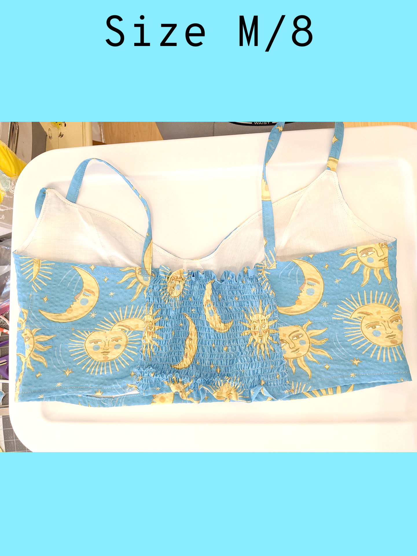 Back view of a light blue cropped tank top with a yellow sun and moon print and gold topstitching, displayed flat on a white surface. Center back includes a rushed or scrunched effect on the fabric. With a text overlay that reads, "Size M/8".