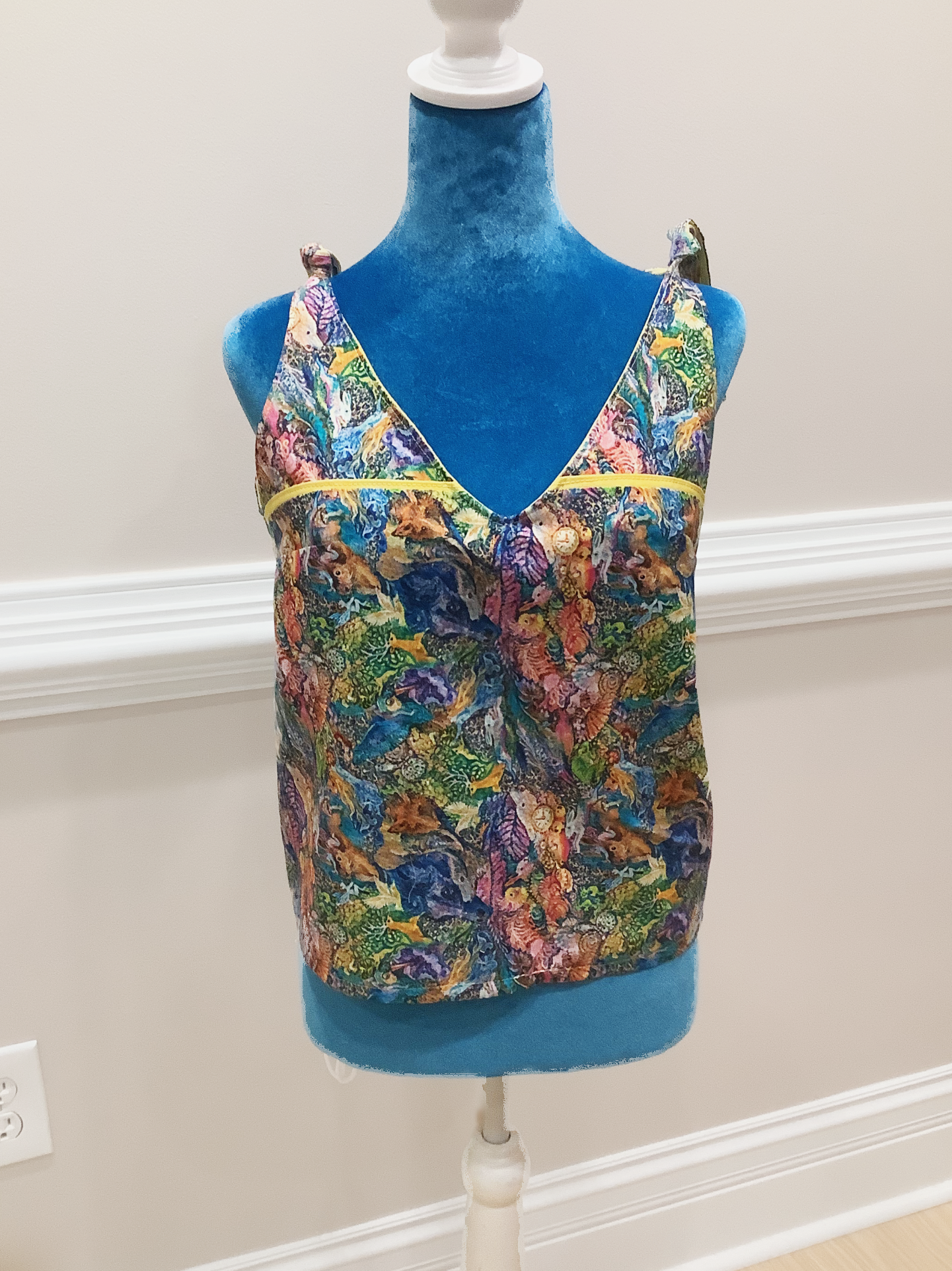 Tank top with a bright animal pattern. Has ties on the shoulders and is on a blue manniquin in a beige background.