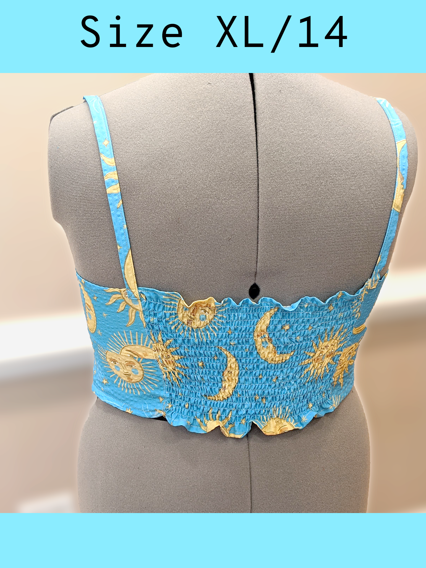 Back view of a light blue cropped tank top with a yellow sun and moon print and gold topstitching, displayed on a mannequin. Center back features a gathered, or rushed effect for a tighter fit. With a text overlay that reads, "Size XL/14".