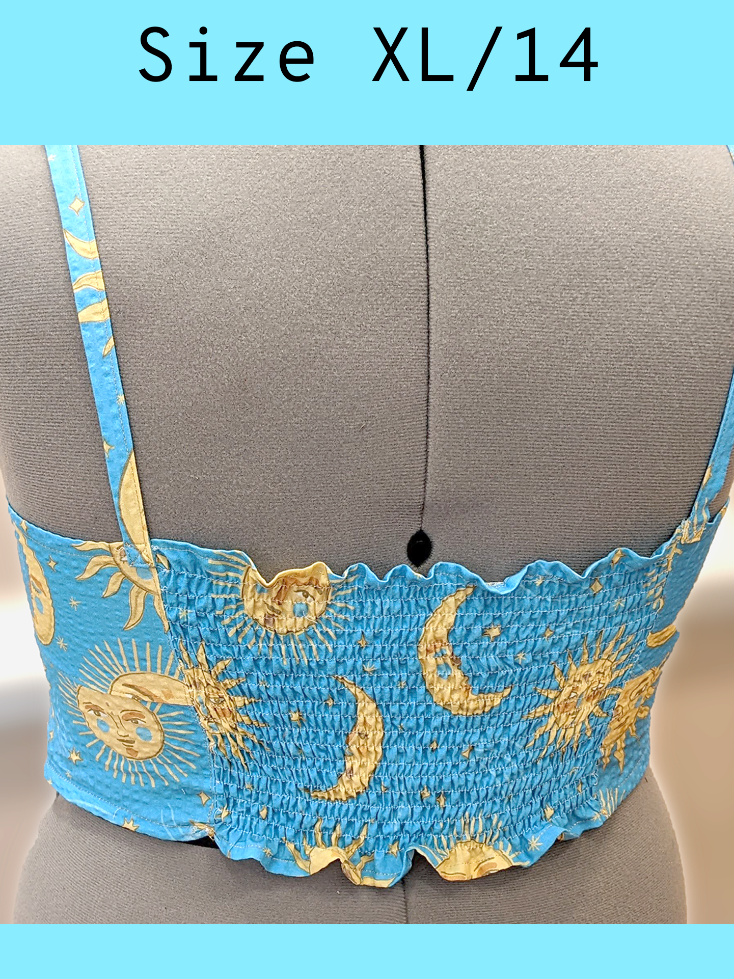Zoomed in back view of a light blue cropped tank top with a yellow sun and moon print and gold topstitching, displayed on a mannequin. Center back features a gathered, or rushed effect for a tighter fit. With a text overlay that reads, "Size XL/14".
