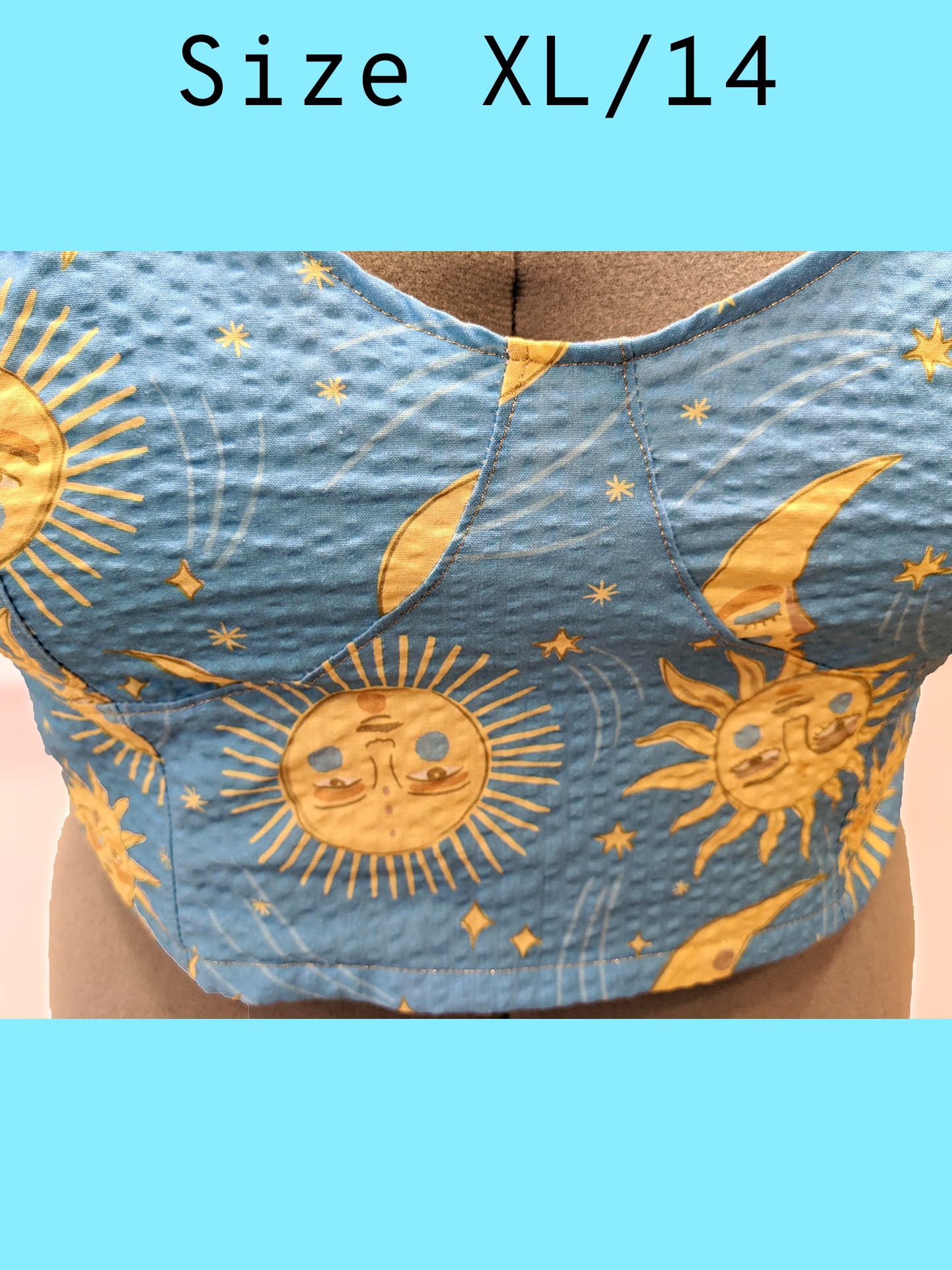 Close up of the front of a light blue cropped tank top with a yellow sun and moon print and gold topstitching, displayed on a mannequin. With a text overlay that reads, "Size XL/14".