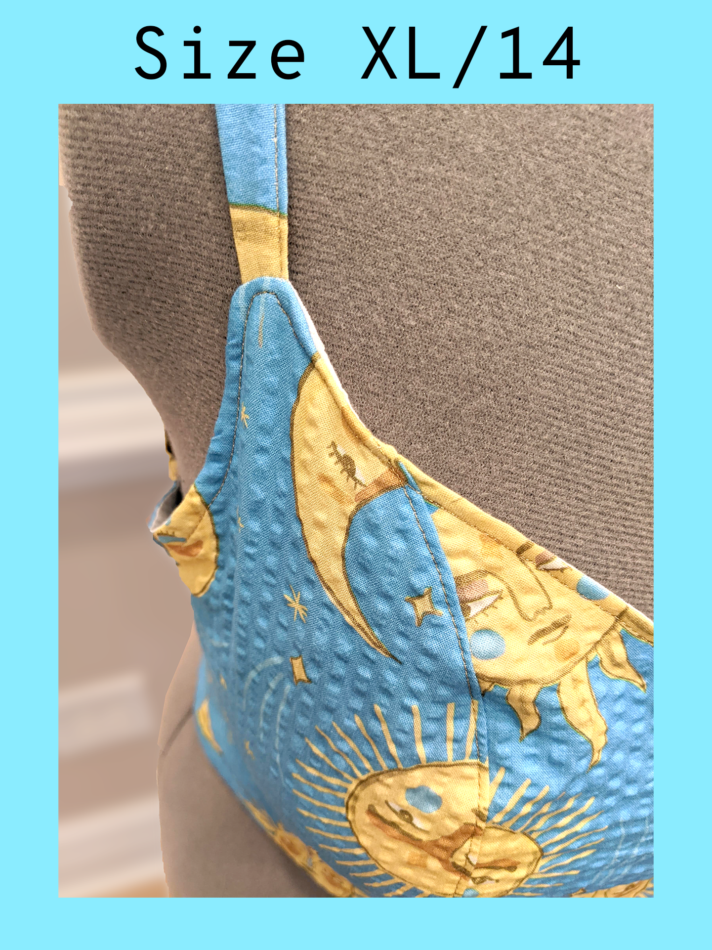 Close up of the front strap of a light blue cropped tank top with a yellow sun and moon print and gold topstitching, displayed on a mannequin. With a text overlay that reads, "Size XL/14".